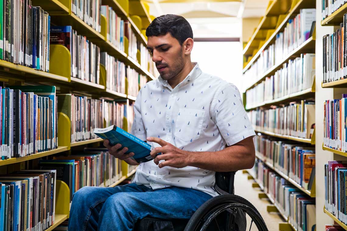 Man in a wheelchair, inside the book stacks, reading a book.